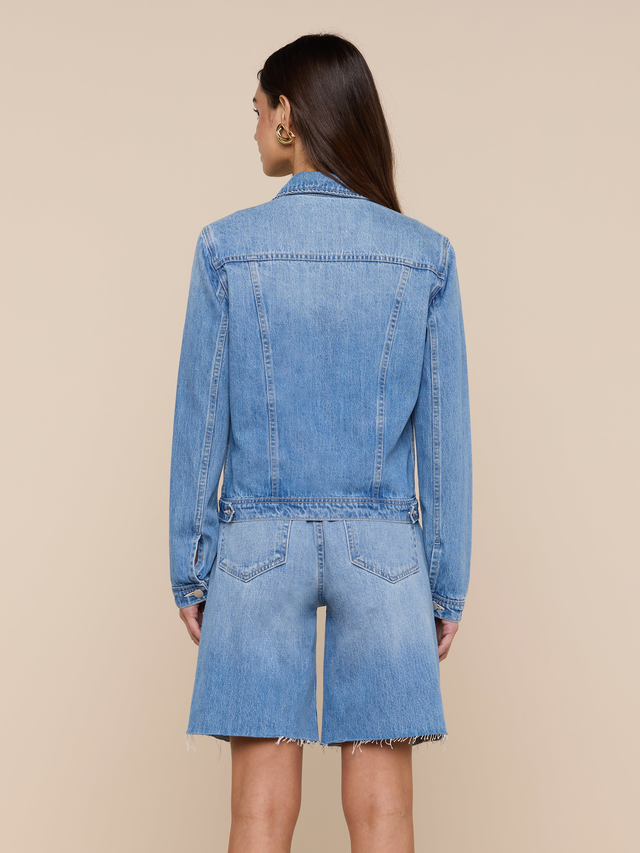 A woman with long brown hair stands facing away, wearing a slim fit Celine Jean Jacket by L’AGENCE in a light denim wash and matching knee-length denim shorts with frayed hems against a plain light background.