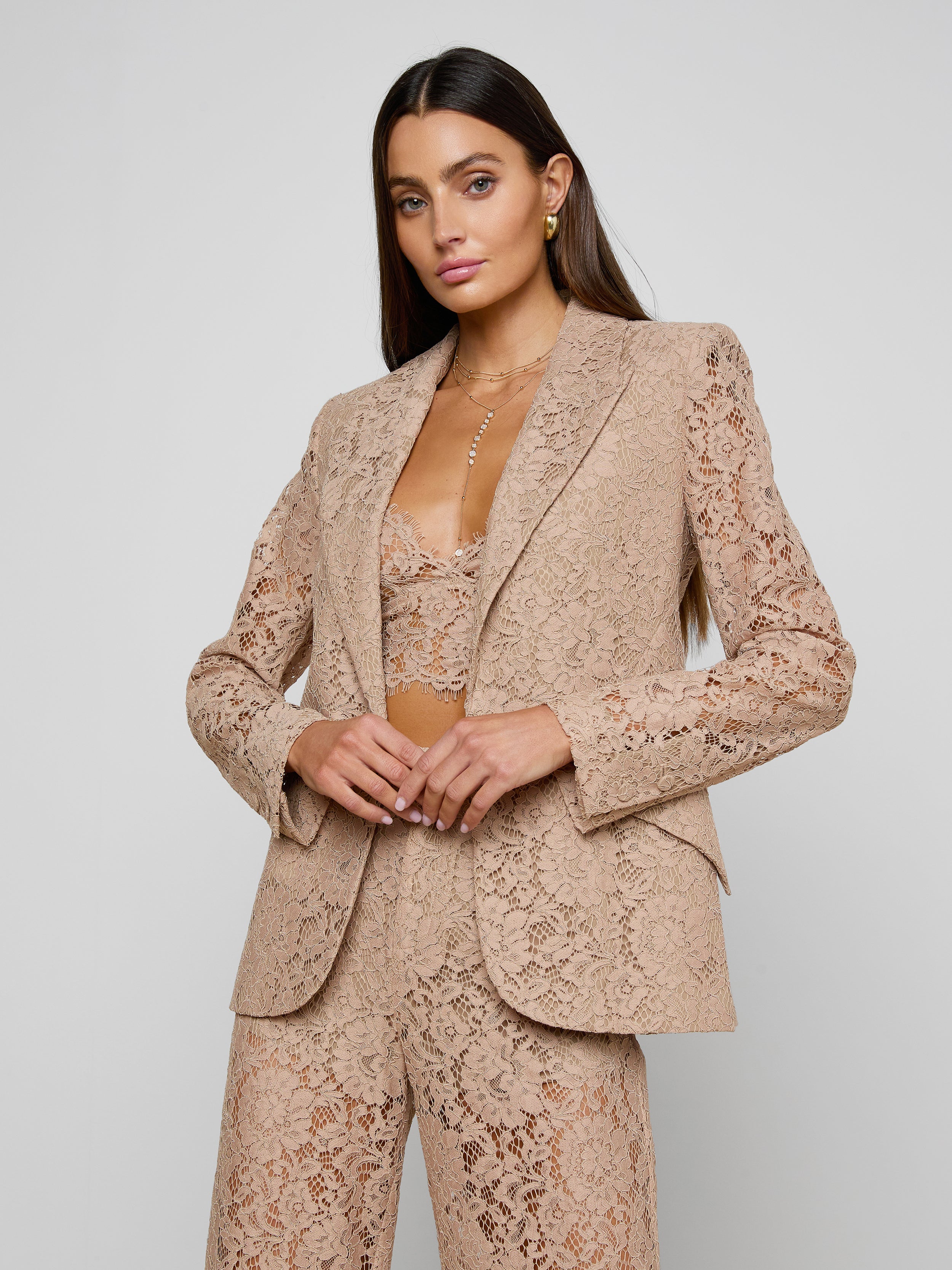 A woman models the Chamberlain Lace Blazer with a matching tan lace bralette underneath, standing against a plain light background and posing with her hands at her waist for an office-ready style.