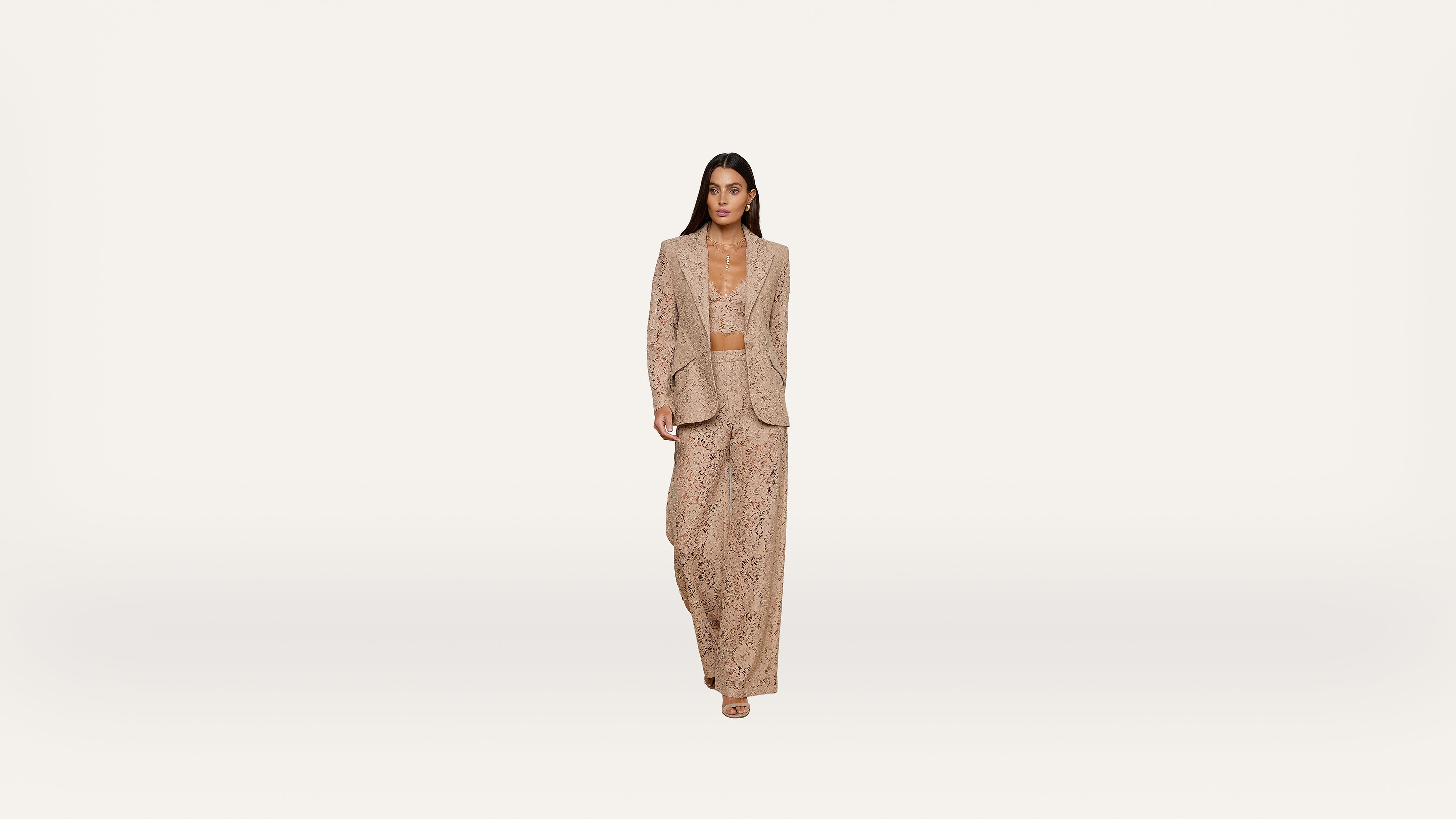 A woman wearing a beige, patterned pantsuit and heels walks confidently toward the camera against a plain white background.