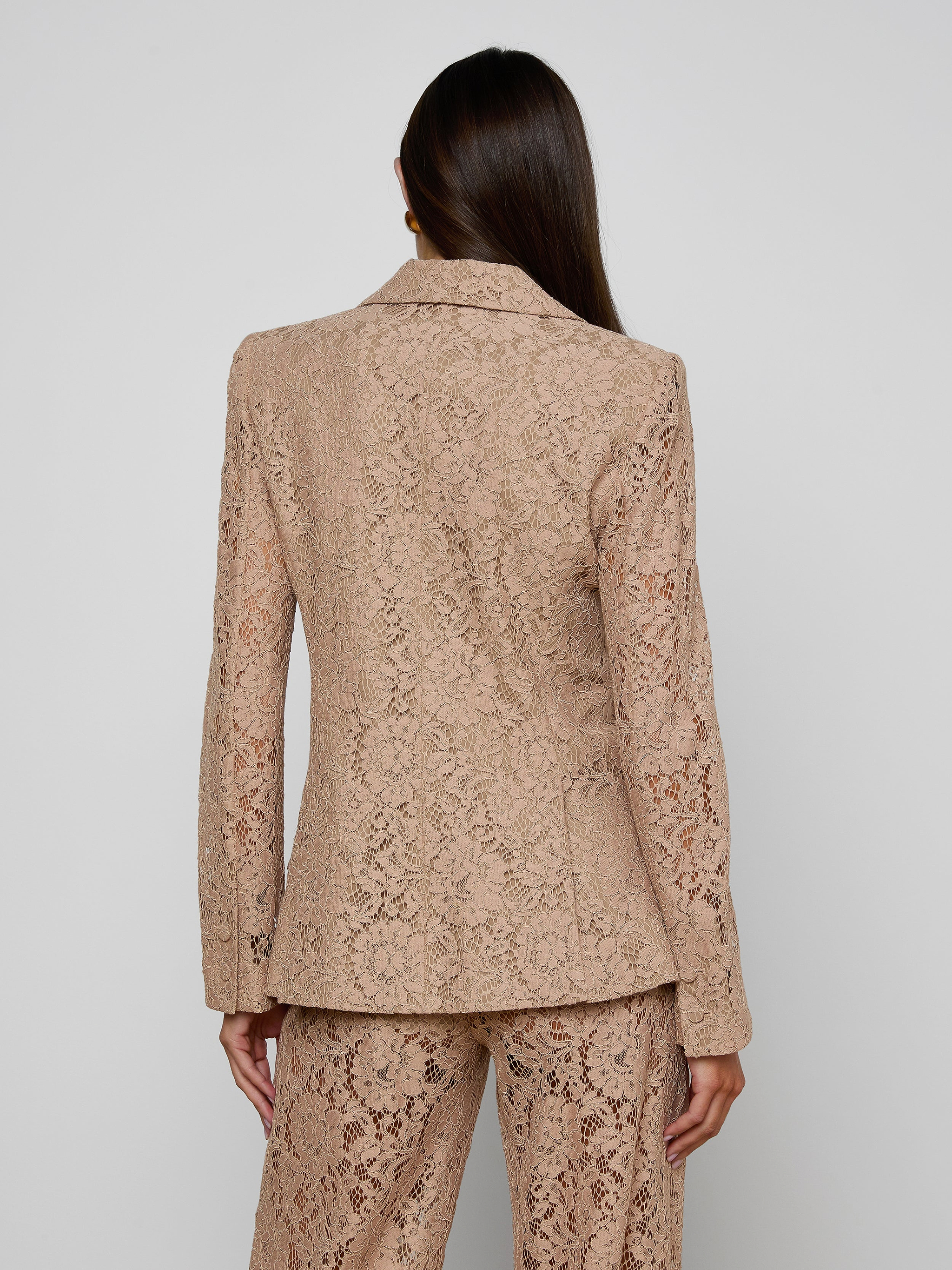 A woman with long dark hair stands with her back to the camera against a plain light background, modeling the Chamberlain Lace Blazer and matching pants.