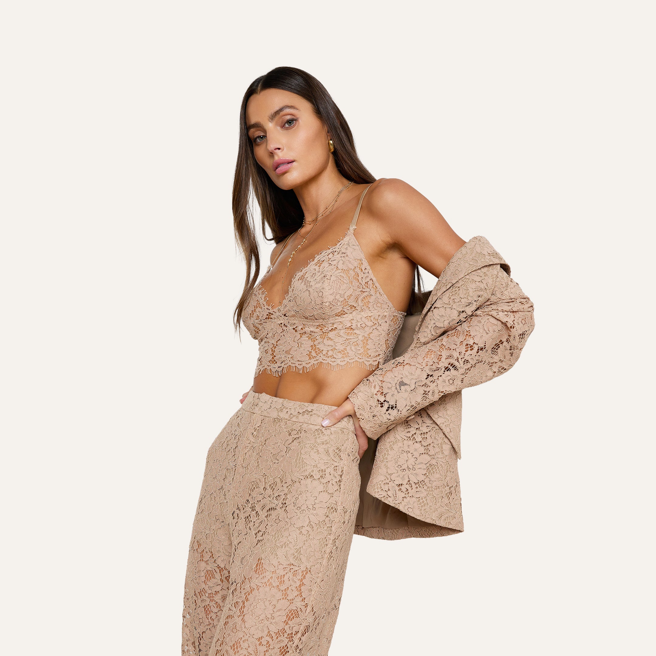 A woman poses against a plain background wearing the Chamberlain Lace Blazer draped over one shoulder, styled with a beige lace bralette and high-waisted skirt.
