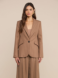 Una donna dai lunghi capelli castani indossa il blazer Chamberlain a righe sottili color cuoio, abbinato a pantaloni da completo leggeri a gamba larga, in piedi davanti a uno sfondo beige a tinta unita.