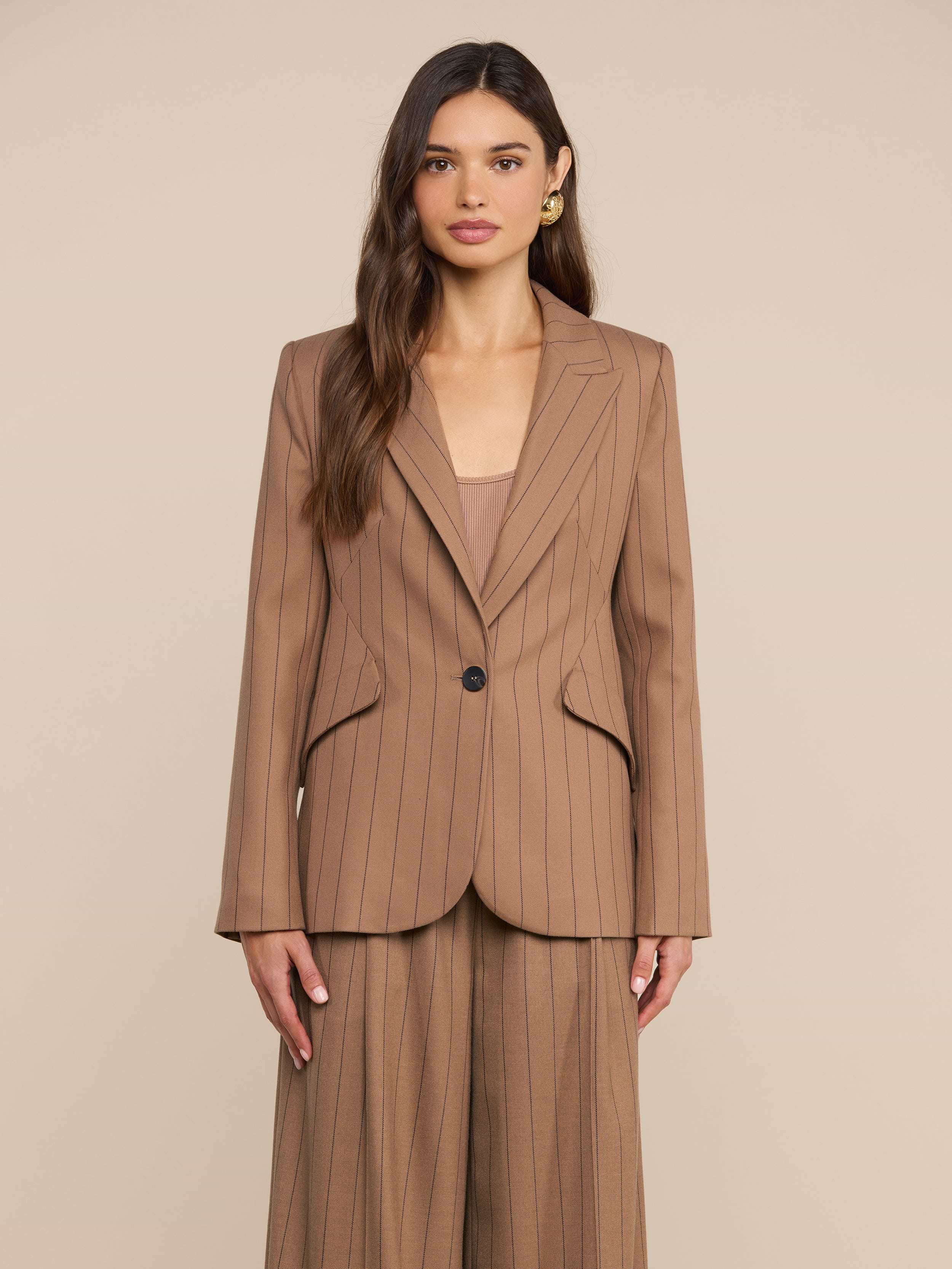 Una donna dai lunghi capelli castani indossa il blazer Chamberlain a righe sottili color cuoio, abbinato a pantaloni da completo leggeri a gamba larga, in piedi davanti a uno sfondo beige a tinta unita.