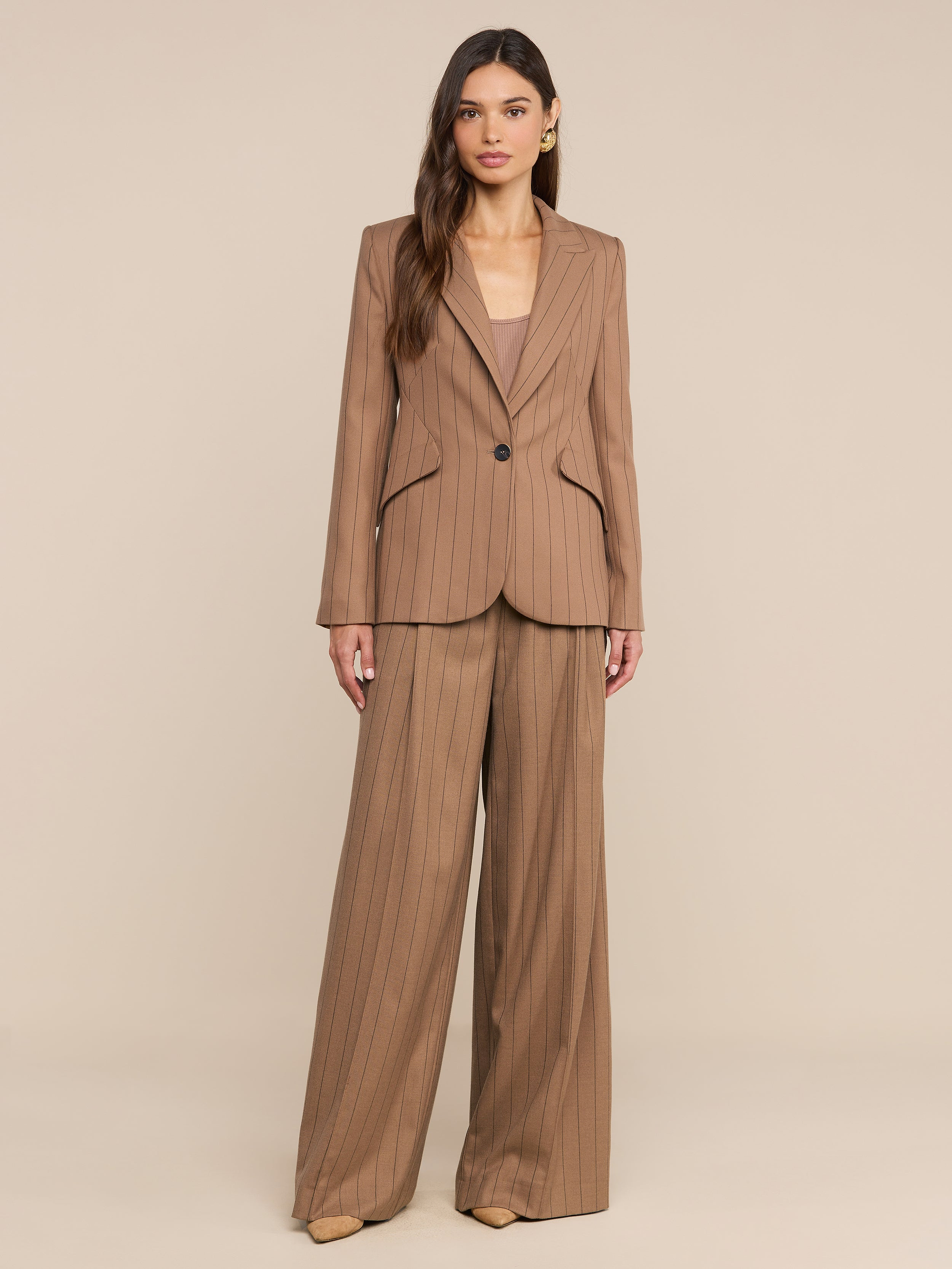 Una donna è ritratta di fronte, con indosso il Chamberlain Blazer: una giacca color cuoio a righe sottili abbinata a pantaloni da tailleur a gamba larga e un top beige. Ha lunghi capelli castani, indossa tacchi di colore neutro e si trova in piedi davanti a uno sfondo beige a tinta unita.