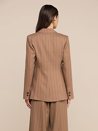 Una donna dai lunghi capelli castani è in piedi su uno sfondo beige e indossa il blazer Chamberlain a righe sottili color cuoio, a maniche lunghe, abbinato a pantaloni a gamba larga dello stesso tessuto, realizzati in un leggero tessuto da completo. È ritratta di schiena.