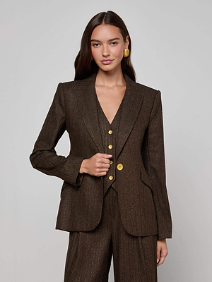 A woman with long brown hair models the Chamberlain Pinstriped Blazer by L'AGENCE—featuring gold buttons—paired with statement gold earrings, set against a plain light gray background.