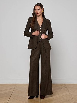 A woman stands indoors against a plain light-colored wall, wearing the L'AGENCE Chamberlain Pinstriped Blazer with wide-leg trousers. Her outfit is paired with pointed-toe shoes, loose hair, and gold earrings.