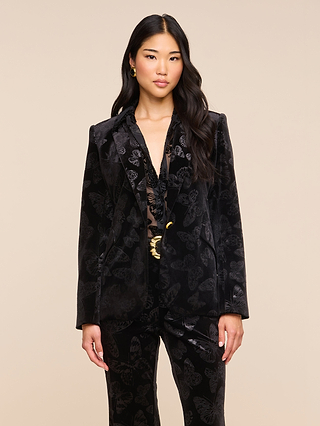 A woman with long black hair wears the Chamberlain Velvet Blazer over a lace top, paired with gold earrings, standing against a plain beige background.
