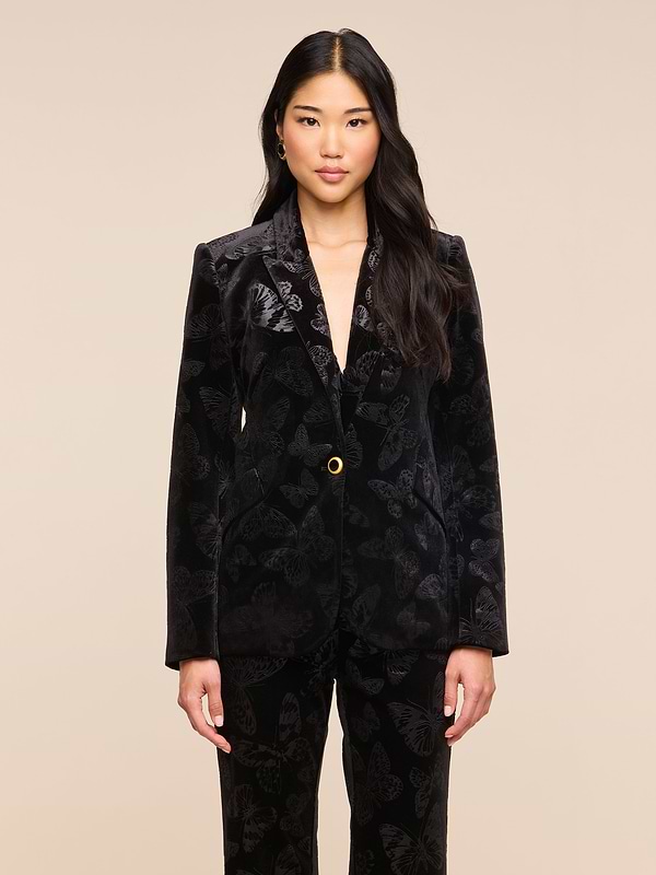 A woman with long dark hair wears the Chamberlain Velvet Blazer by L’AGENCE and matching pants, standing against a plain light background, facing forward with her arms relaxed at her sides.