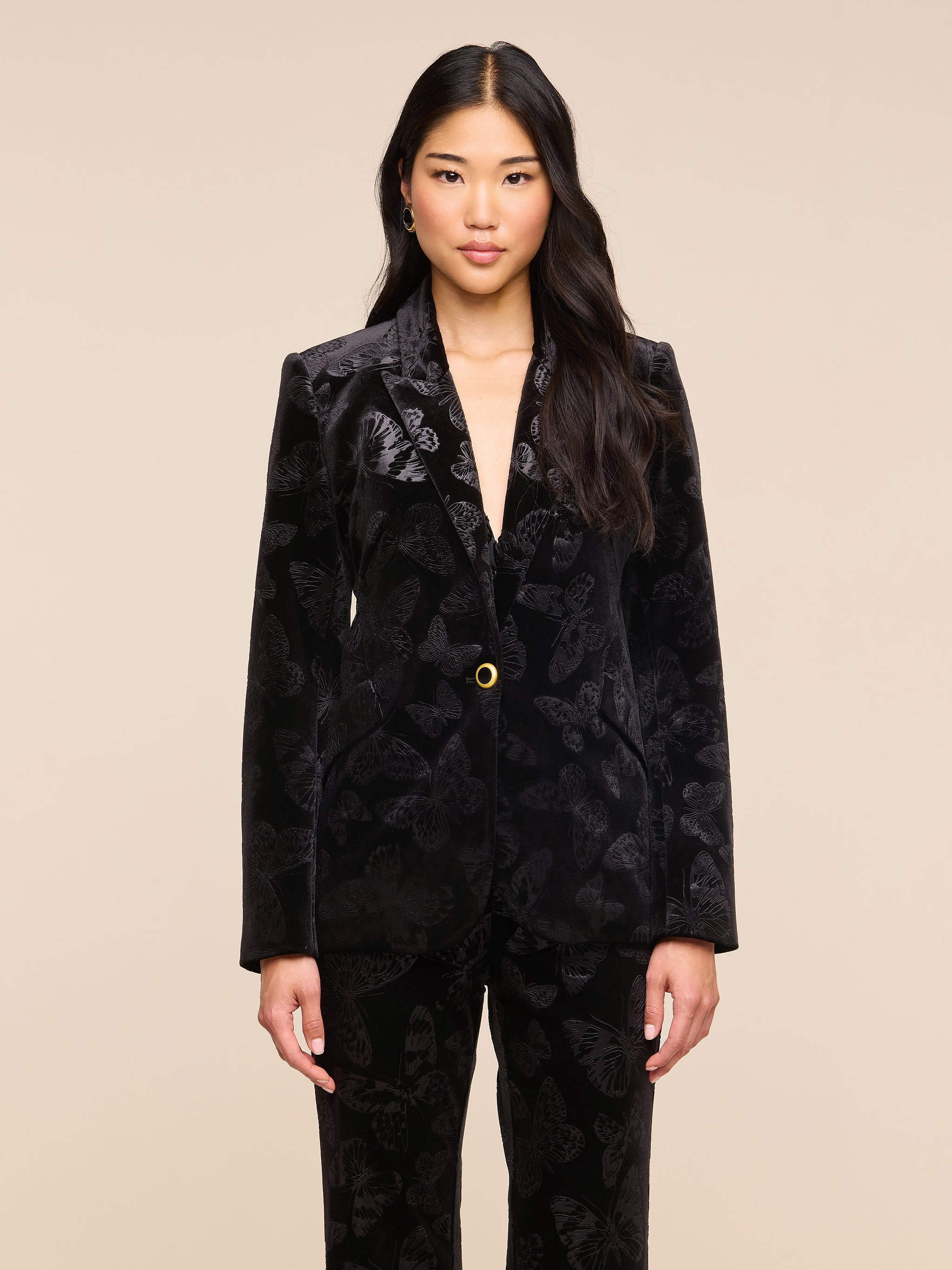 A woman with long dark hair wears the Chamberlain Velvet Blazer by L’AGENCE and matching pants, standing against a plain light background, facing forward with her arms relaxed at her sides.