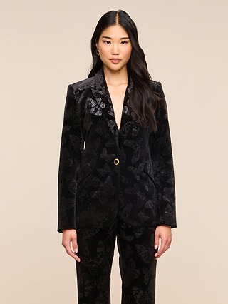 A woman stands facing forward, wearing the Chamberlain Velvet Blazer by L’AGENCE, which features a butterfly-embossed design and a single button closure. She has long, wavy black hair and maintains a neutral expression. The background is plain beige.