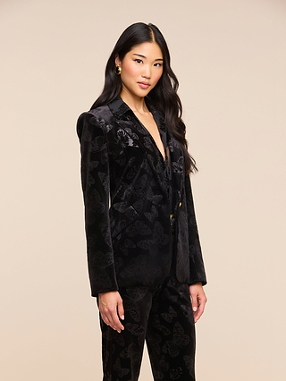 A woman with long black hair wears the Chamberlain Velvet Blazer, featuring a butterfly-embossed design, as she stands against a plain beige background looking at the camera.