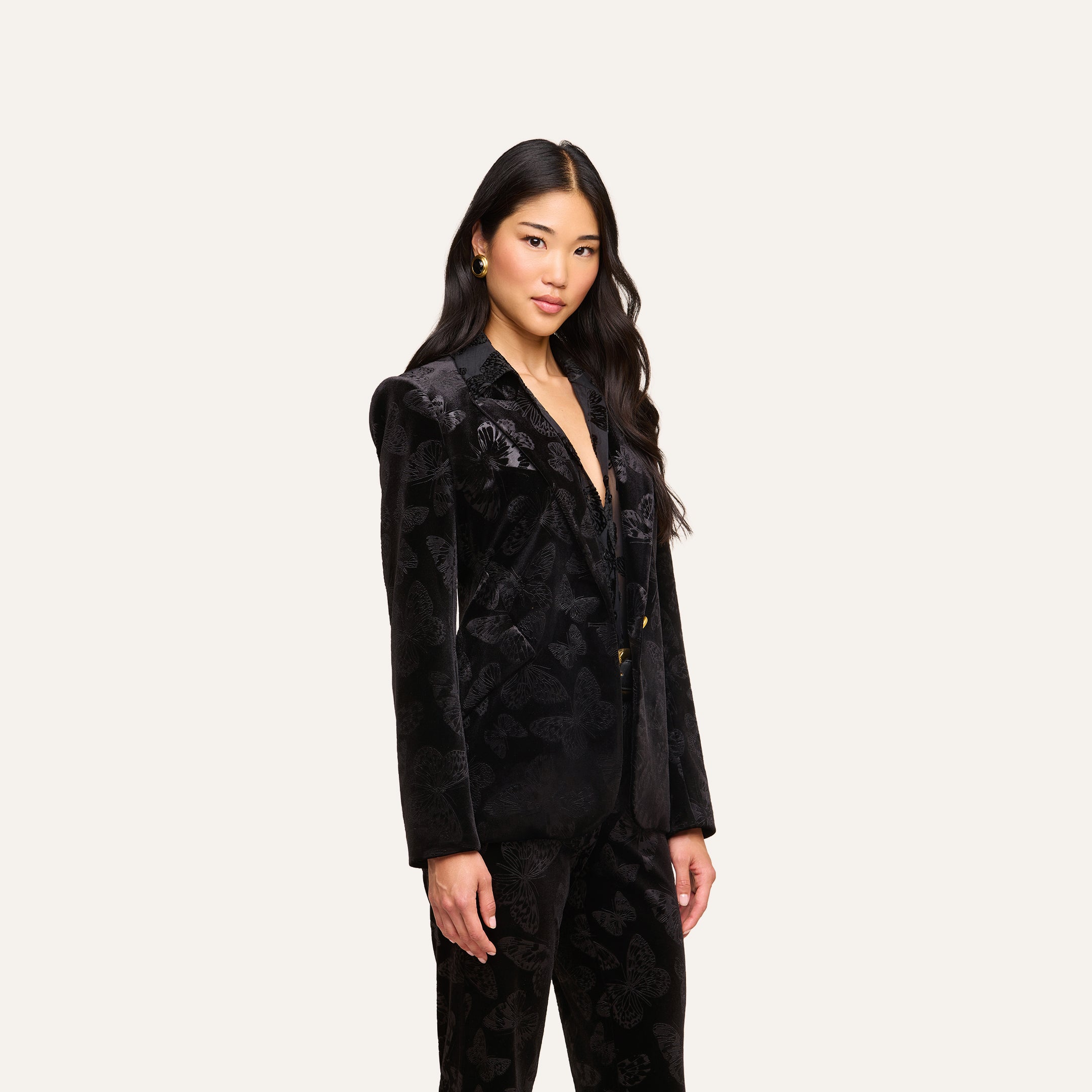 A woman with long dark hair stands against a plain white background, looking slightly to the side with a neutral expression. She is wearing the L’AGENCE Chamberlain Velvet Blazer and matching pants.