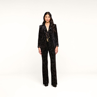 A woman stands against a plain white background, wearing the Chamberlain Velvet Blazer with flared pants. She has long, wavy hair and is looking straight ahead with a neutral expression.