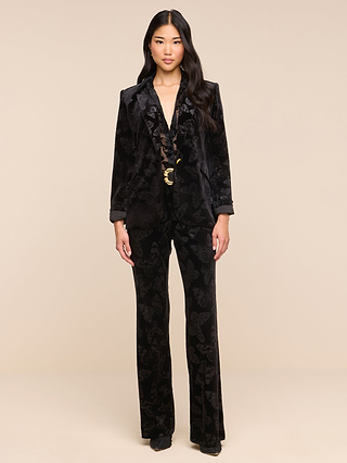 A woman stands facing forward, wearing the Chamberlain Velvet Blazer embossed with butterflies and its matching suit. She pairs the look with black heels and a black blouse, cinched at the waist with a gold belt. Her long, wavy black hair frames a neutral expression as she poses against a beige background, effortlessly showcasing the timeless elegance of the Chamberlain Velvet Blazer.