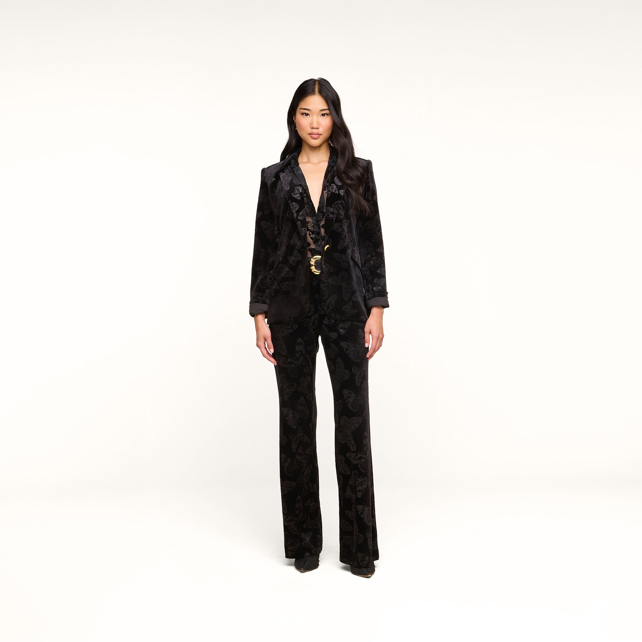 A woman stands against a plain white background, wearing the Chamberlain Velvet Blazer with flared pants. She has long, wavy hair and is looking straight ahead with a neutral expression.