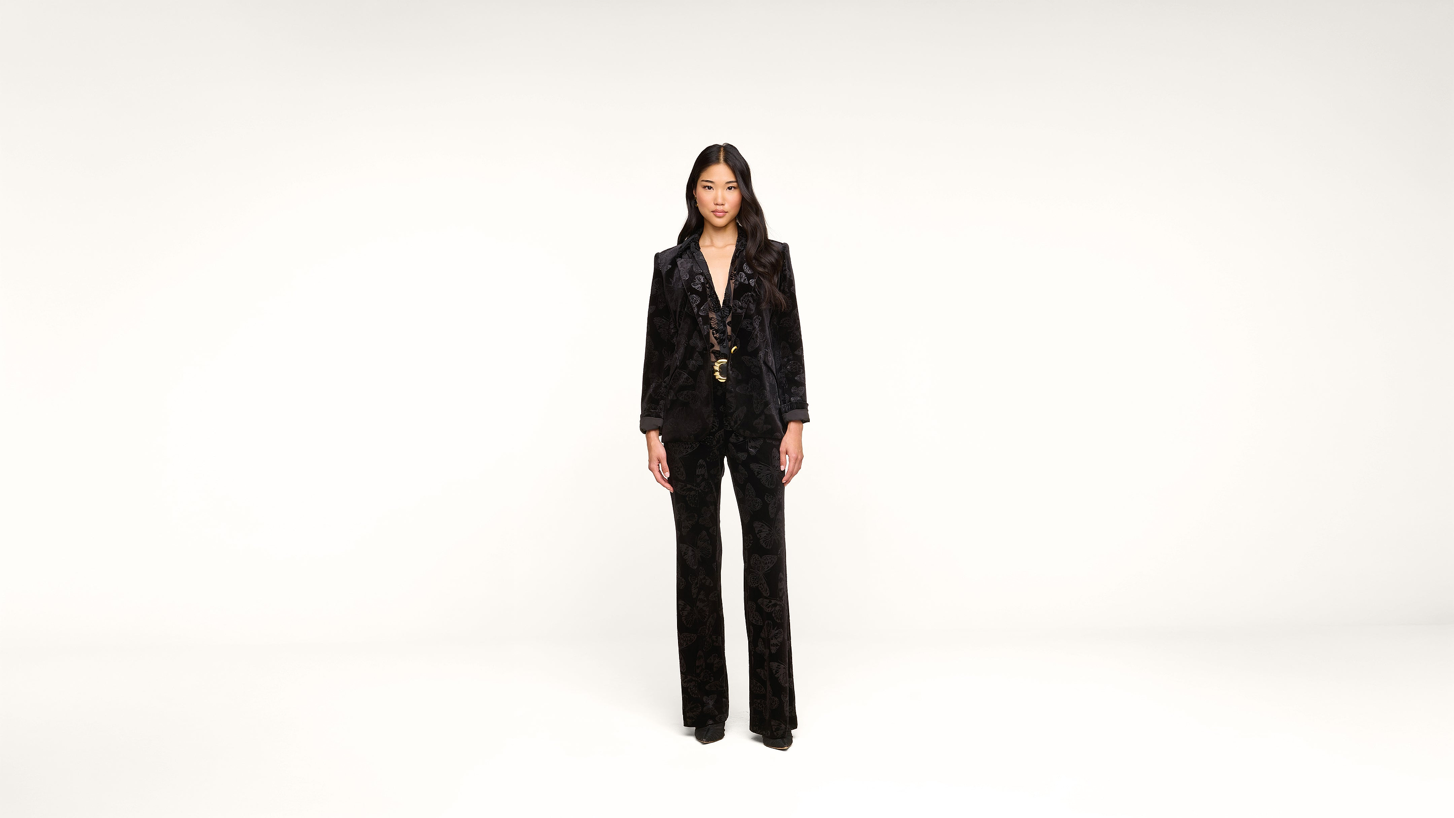 A woman stands facing forward in a black velvet suit and heels against a plain white background.