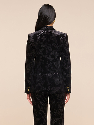 A person with long dark hair, seen from behind, wears the Chamberlain Velvet Blazer—fitted and black with butterfly embossing and gold cuff buttons—along with matching pants featuring subtle butterfly patterns, set against a plain beige background.