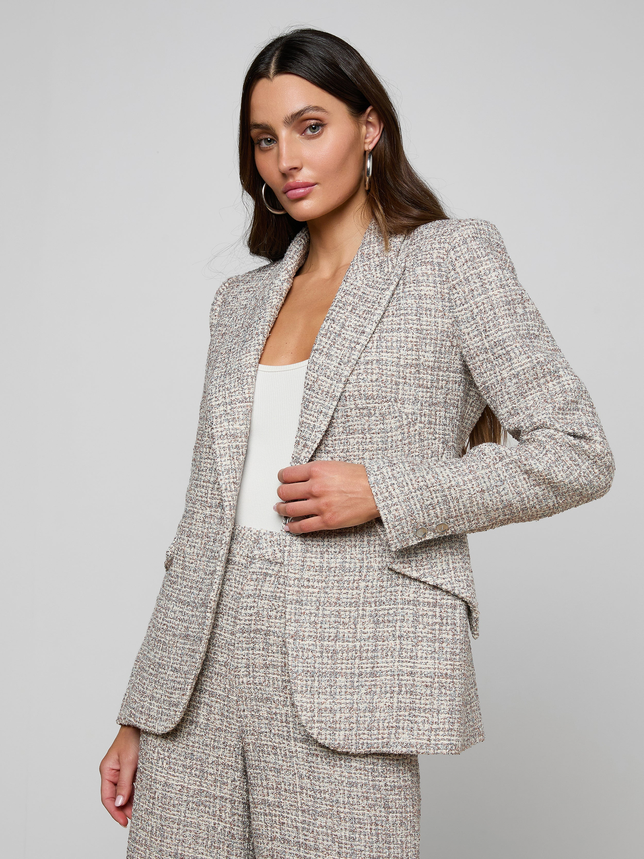 Wearing the Chamberlain Tweed Blazer with matching trousers and a white top underneath, a woman with long dark hair and hoop earrings poses against a plain, light background.