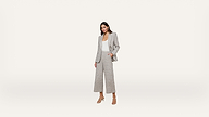 A woman stands against a plain white background, wearing a light gray suit with wide-leg pants, a white top, and beige heeled sandals, with one hand in her pocket.