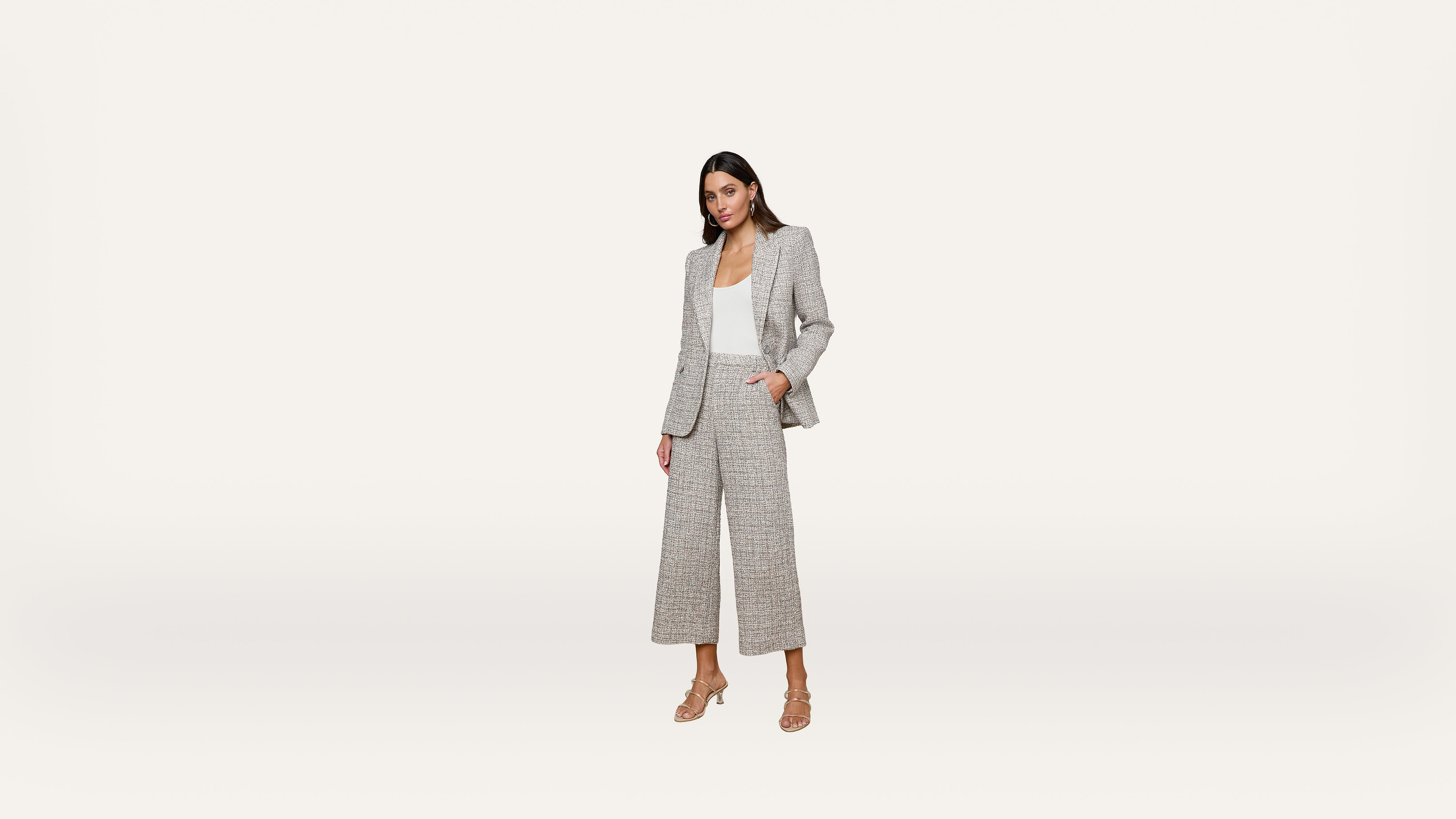 A woman stands against a plain white background, wearing a light gray suit with wide-leg pants, a white top, and beige heeled sandals, with one hand in her pocket.