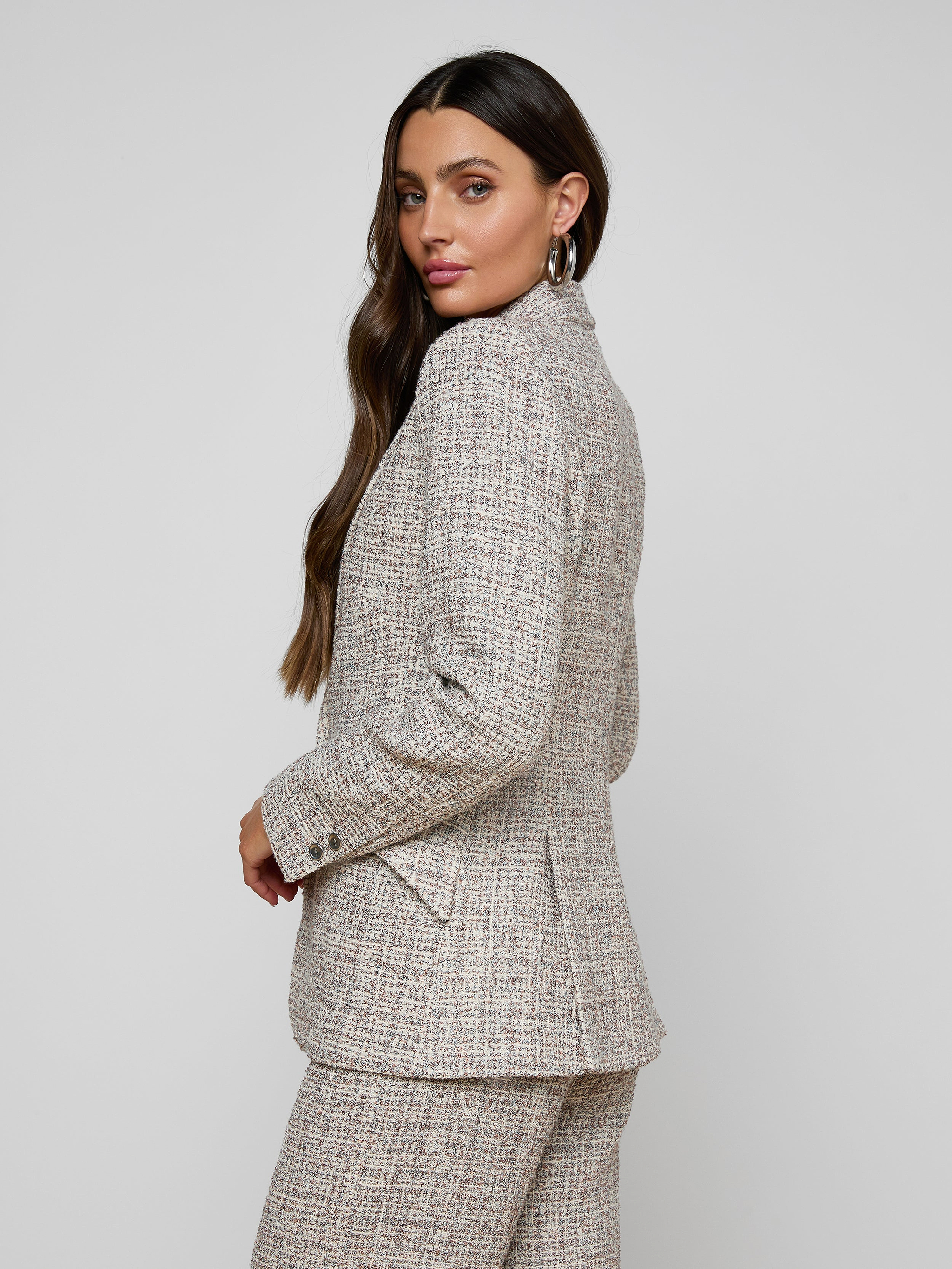 A woman with long brown hair models the Chamberlain Tweed Blazer, standing slightly turned to the side and looking over her shoulder against a plain, light background.