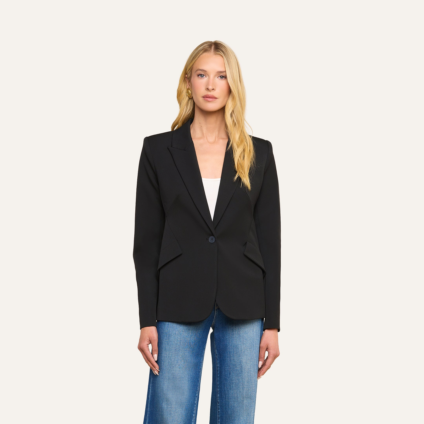A woman with long blonde hair wears the Chamberlain Blazer, a precision-tailored black single-breasted jacket, over a white top and blue jeans. She stands against a plain beige background, looking at the camera with a neutral expression.