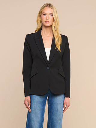A woman with long blonde hair wears the Chamberlain Blazer, a precision-tailored black single-breasted jacket, over a white top and blue jeans. She stands against a plain beige background, looking at the camera with a neutral expression.
