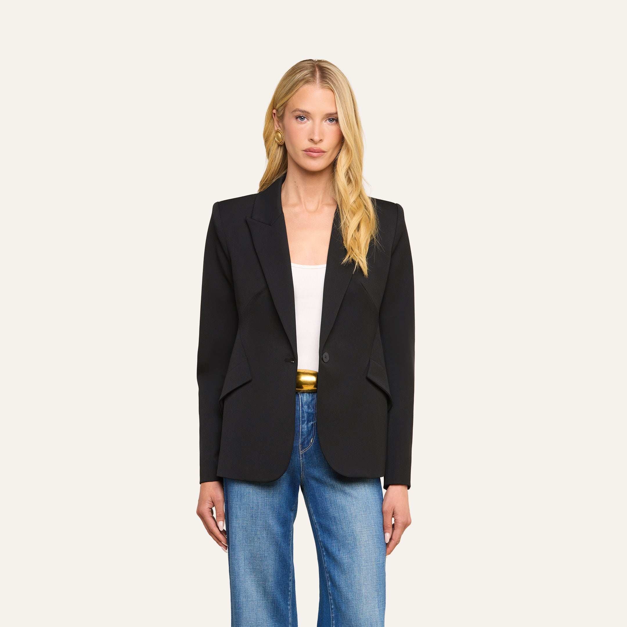 A woman with long blonde hair, wearing the Chamberlain Blazer—crafted from black twill suiting in a single-breasted silhouette—over a white top and blue jeans, stands facing forward against a plain light background.