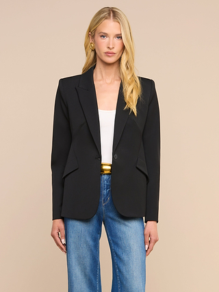 A woman with long blonde hair wears the Chamberlain Blazer, a single-breasted black twill suiting jacket, paired with a white top and blue jeans. She stands against a plain beige background, looking straight at the camera with a neutral expression. The blazer’s precision tailoring adds structure and sophistication to her timeless look.