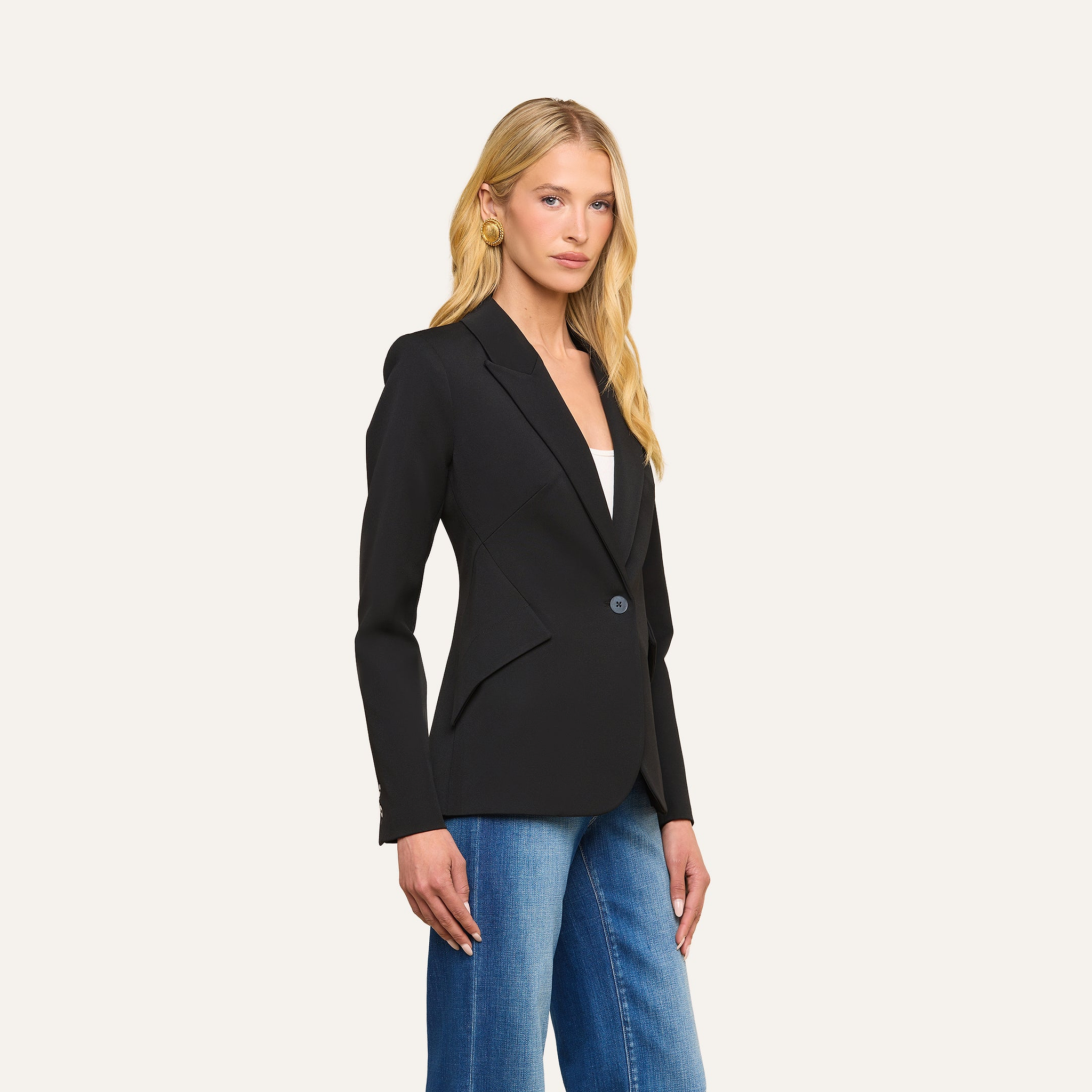 A woman with long blonde hair wears the Chamberlain Blazer—a fitted, single-breasted jacket crafted from black twill suiting with precision tailoring—over a white top and blue jeans, standing against a plain light background.