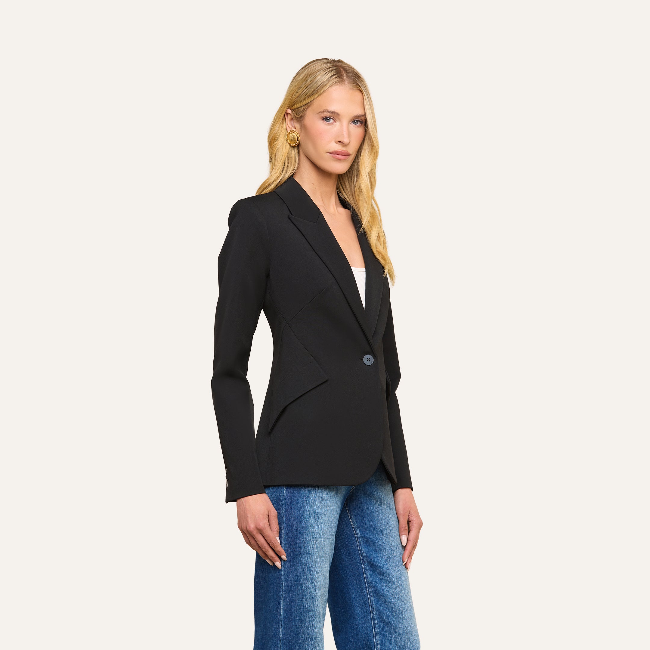 A woman with long blonde hair wears the Chamberlain Blazer—a fitted, single-breasted jacket crafted from black twill suiting with precision tailoring—over a white top and blue jeans, standing against a plain light background.