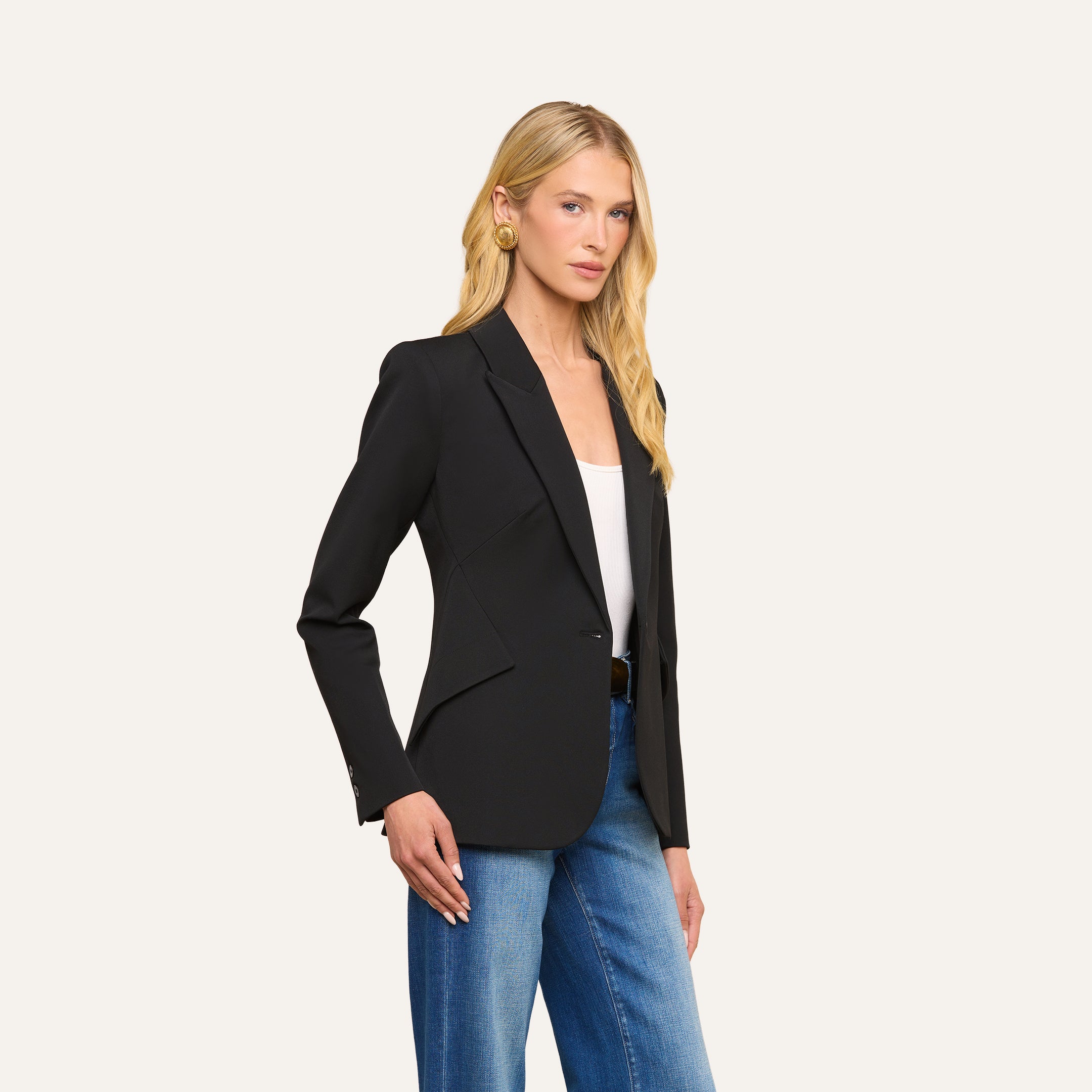 A woman with long blonde hair wears the Chamberlain Blazer—a black twill suiting single-breasted jacket—over a white top and blue jeans, standing against a plain light background facing forward with one hand at her side.