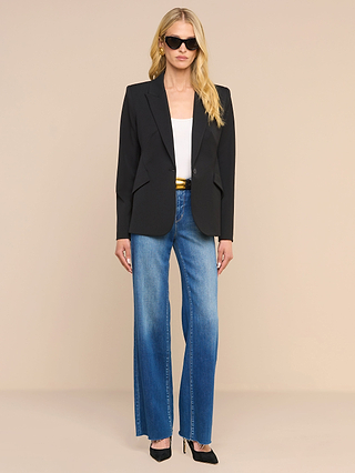 A woman wearing sunglasses, a white top, blue jeans, black heels, and the Chamberlain Blazer—crafted in black twill suiting with precision tailoring—stands against a plain beige background.