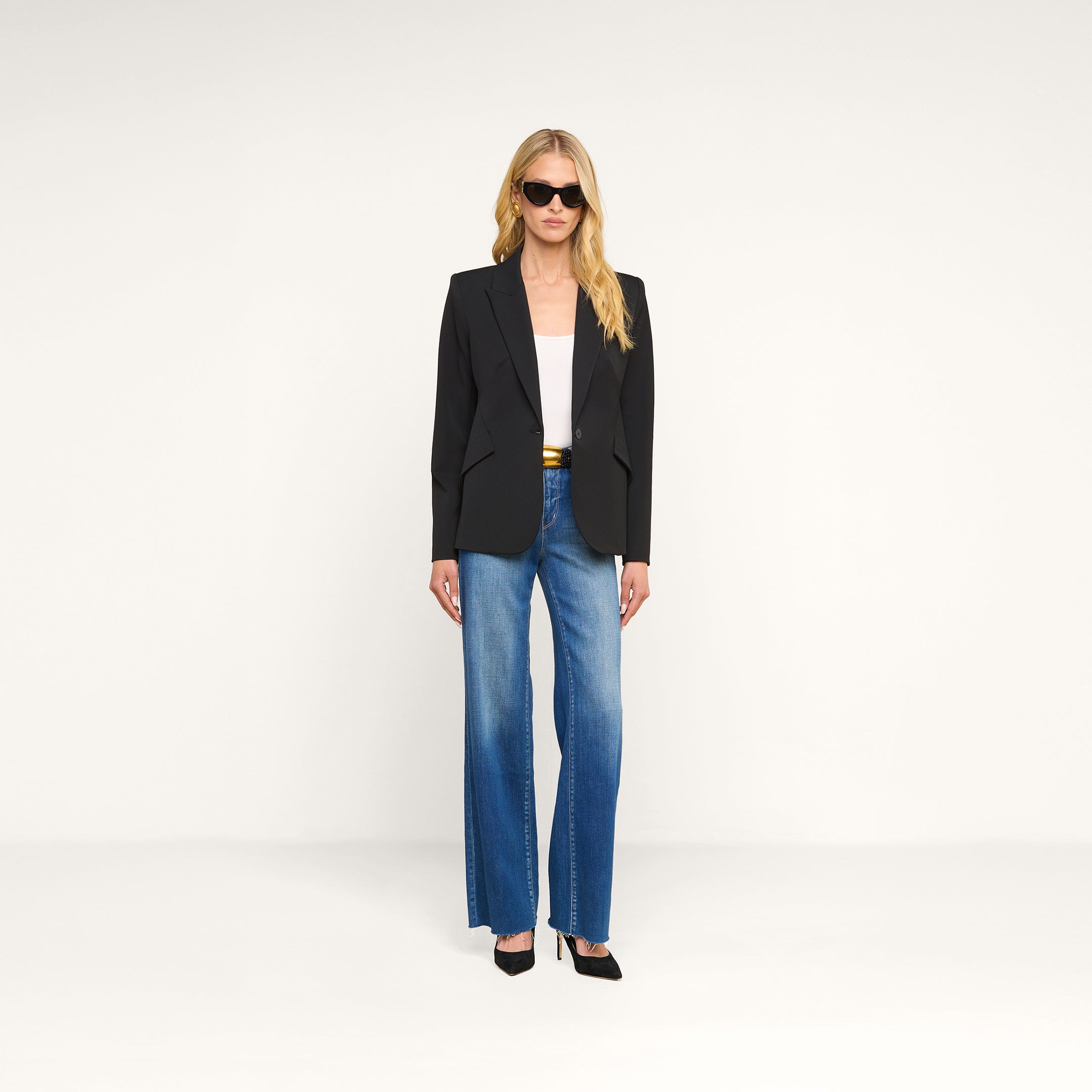 A person stands against a plain white background, facing forward with a neutral expression, wearing sunglasses and the Chamberlain Blazer—a black single-breasted blazer with precision tailoring—over a white top, paired with blue wide-leg jeans and black heels.