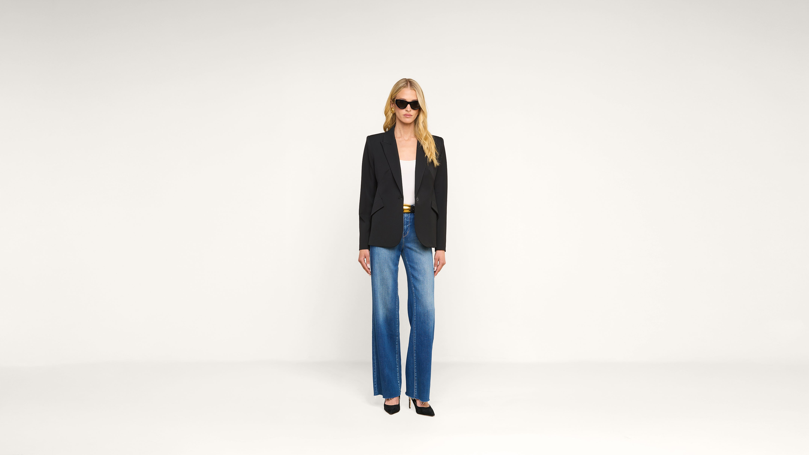 A person stands against a plain white background, wearing a black blazer, white top, blue wide-leg jeans, black heels, and sunglasses.