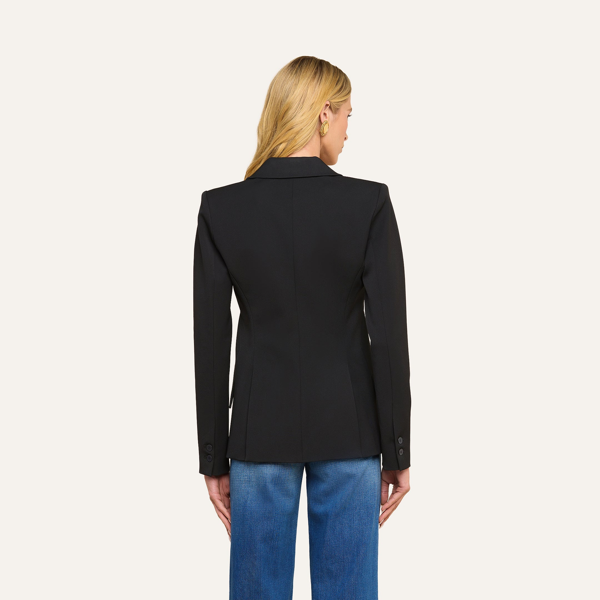 A woman with long blonde hair is seen from the back, wearing the precision-tailored Chamberlain Blazer and blue jeans, standing against a plain light background.