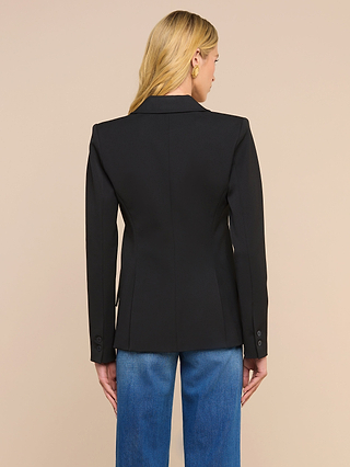 A woman with blonde hair, wearing the Chamberlain Blazer in black twill suiting and blue jeans, stands facing away from the camera against a plain beige background.