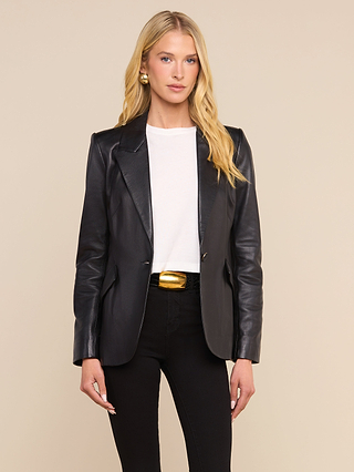 A woman with long blonde hair wears the Chamberlain Leather Blazer over a white shirt, paired with black pants, gold earrings, and a gold-buckled belt, standing against a plain beige background.
