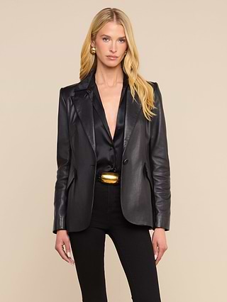 A woman with long blonde hair wears the Chamberlain Leather Blazer, featuring peaked lapels, paired with a black top, black pants, and a gold belt. She stands against a plain, light-colored background, gazing directly at the camera with a neutral expression.