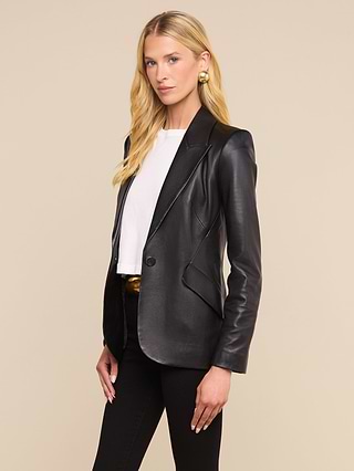 A woman with long blonde hair stands against a plain, light background wearing the Chamberlain Leather Blazer—a black leather blazer with peaked lapels—over a white top and black pants.
