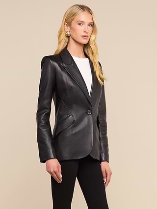 A woman wearing the Chamberlain Leather Blazer, featuring a classic design and peaked lapels in black leather, stands against a plain light background. She pairs it with a white top and black pants, her long blonde hair styled down as she gazes slightly to the side.