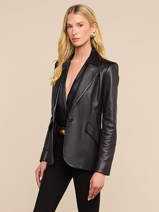 Wearing the Chamberlain Leather Blazer with its lightweight feel and peaked lapels, a woman with long blonde hair stands against a plain light background. She styles it over a low-cut black top and black pants for an effortlessly chic look.