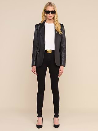 A woman with long blonde hair wears sunglasses, a white shirt, black pants, a gold-buckled belt, black heels, and the Chamberlain Leather Blazer. She stands facing forward against a plain white background.