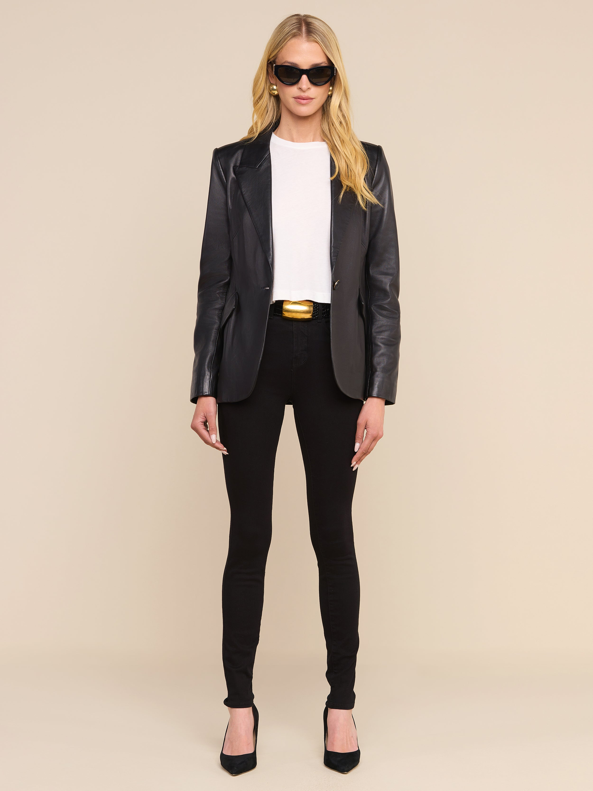 A woman with long blonde hair wears sunglasses, a white shirt, black pants, a gold-buckled belt, black heels, and the Chamberlain Leather Blazer. She stands facing forward against a plain white background.