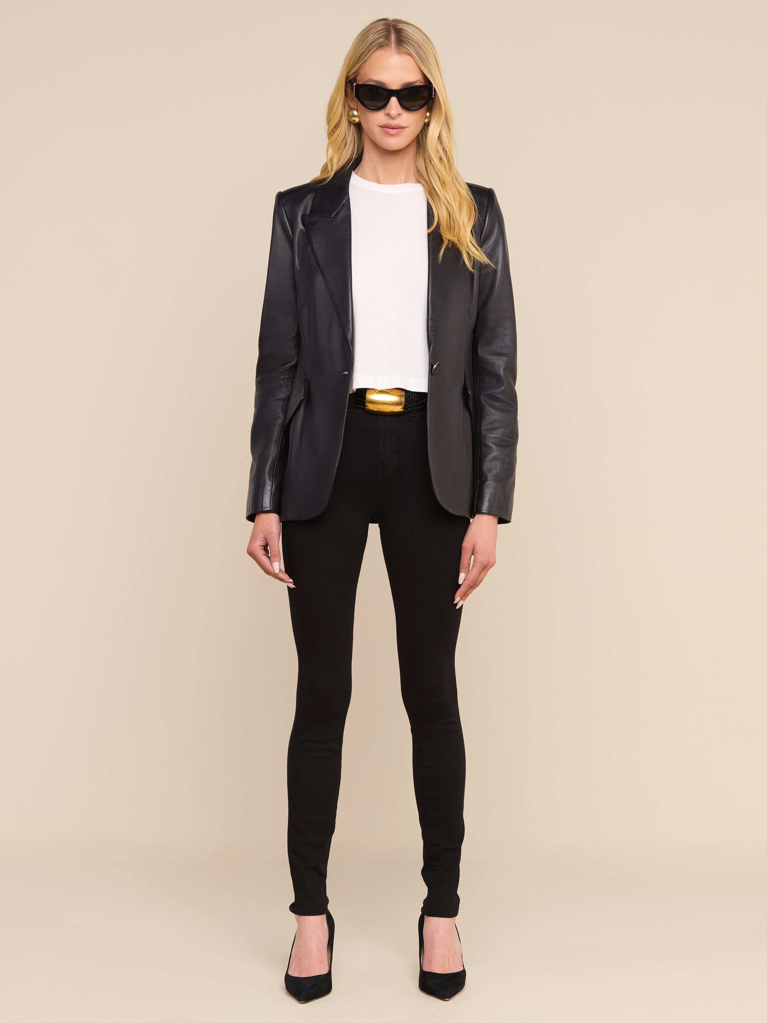 A woman with long blonde hair wears sunglasses, a white shirt, black pants, a gold-buckled belt, black heels, and the Chamberlain Leather Blazer. She stands facing forward against a plain white background.