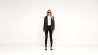 A woman in sunglasses, a black blazer, white top, black pants, and heels stands facing forward against a plain white background.