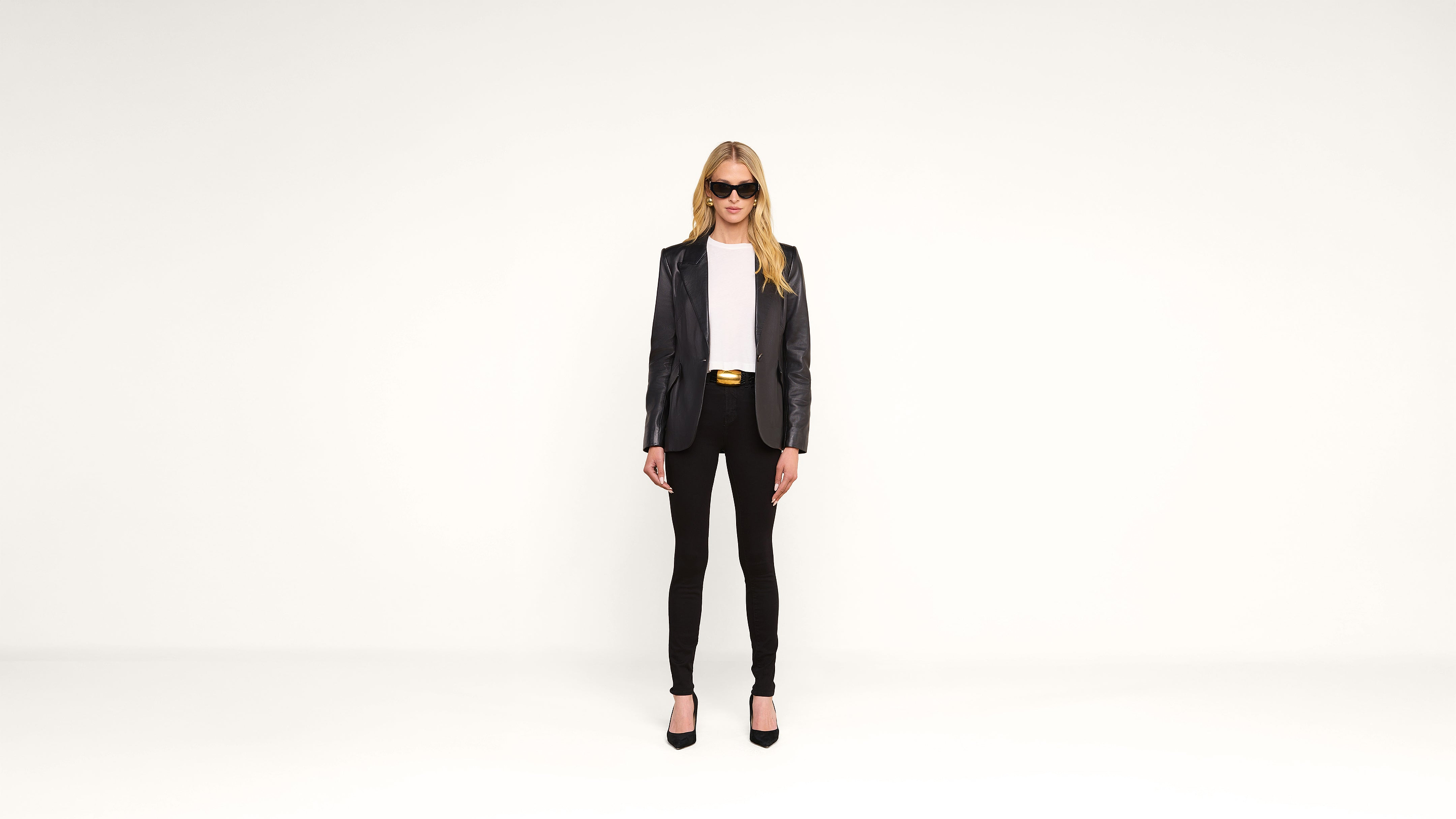 A woman in sunglasses, a black blazer, white top, black pants, and heels stands facing forward against a plain white background.