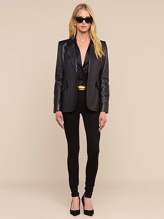 A woman wearing sunglasses and the Chamberlain Leather Blazer—a lightweight black leather piece with a classic design and peaked lapels—paired with black pants and heels, stands against a plain white background, looking forward with her arms relaxed at her sides.