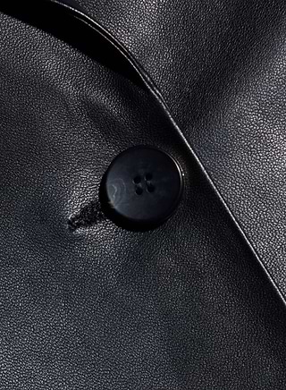 Close-up of a black button sewn onto the shiny black leather fabric of the Chamberlain Leather Blazer, highlighting the texture and sheen of its lightweight leather and timeless, classic design.